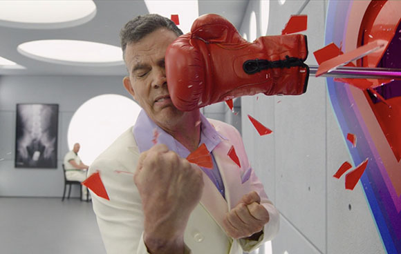 A man in a white suit recoils as a large red boxing glove hits him in the face amidst flying red shards.