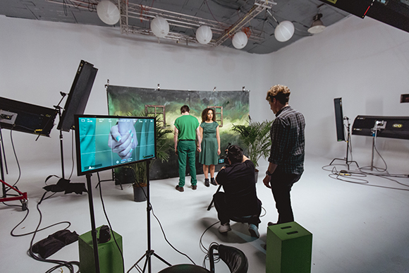 A film crew films two actors standing in front of a green backdrop in a studio.