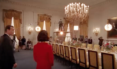 Film crew with lights and cameras in a large, formal dining room with a long table and chandelier.