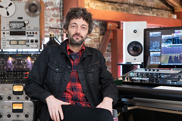 A man with curly hair and a beard sits in a music studio with speakers, monitors, and mixing consoles.