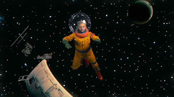 An astronaut in a yellow suit floats in a star-filled space near a planet and space debris.