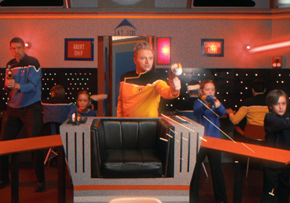 People in colorful sci-fi uniforms posing on a spaceship bridge set with red lighting.