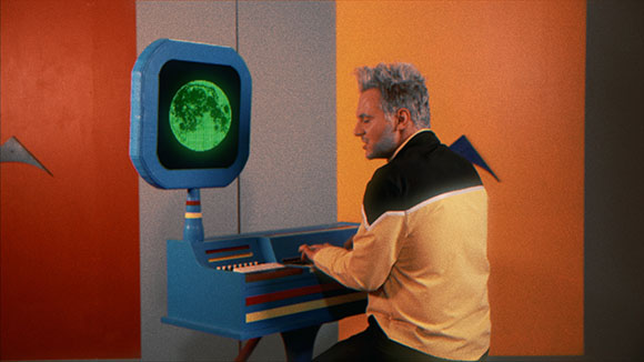 A man in a yellow and black jacket operates a blue futuristic console with a green moon on the screen.