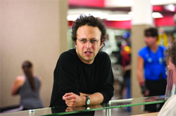 A man with curly hair and glasses wearing a black long-sleeve shirt leans on a glass counter.