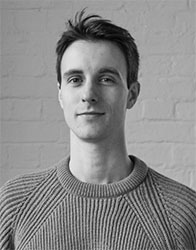 Black and white portrait of a man with short hair wearing a textured ribbed sweater against a brick wall.