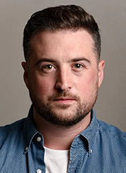 Portrait of a man with short dark hair and a beard wearing a blue button-down shirt.