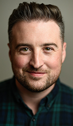 A smiling man with short dark hair, brown eyes, and a light beard wearing a dark plaid shirt.