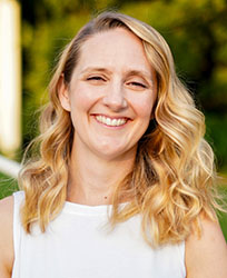 A smiling woman with long, wavy blonde hair wearing a white sleeveless top.