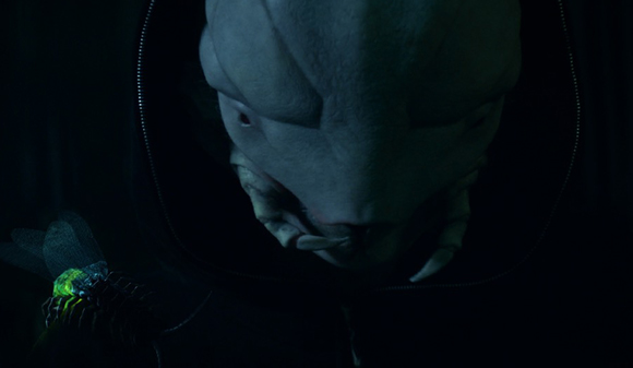 A close-up of a pale, humanoid alien creature with large eyes wearing a dark hooded garment.