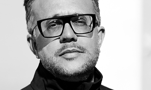 Black and white close-up portrait of a man with a beard and thick black glasses looking forward.