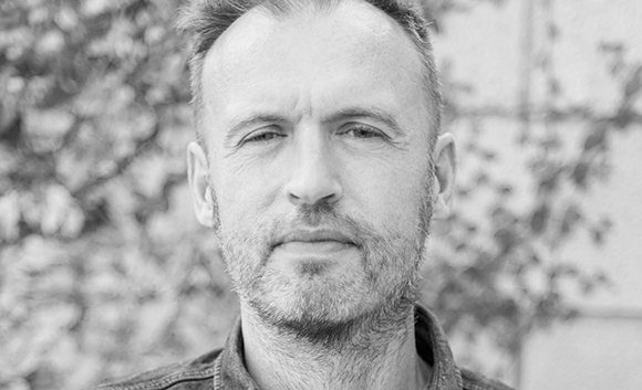 Close-up black and white portrait of a man with short hair and a light beard looking directly at the camera.