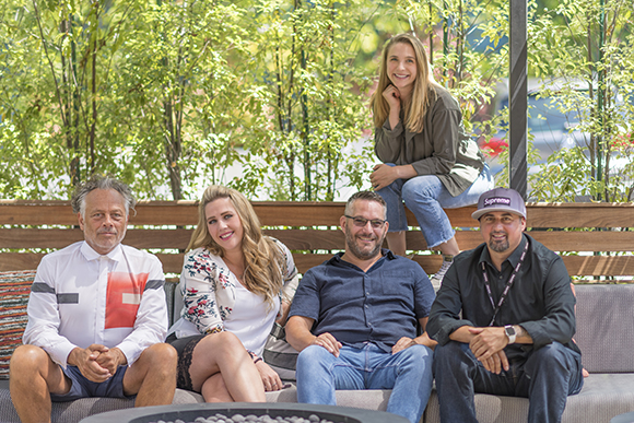 Five smiling people sitting and leaning on a wooden bench in front of green foliage.