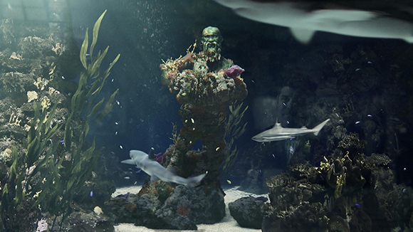 A moss-covered human statue stands underwater amidst seaweed, rocks, and swimming sharks.