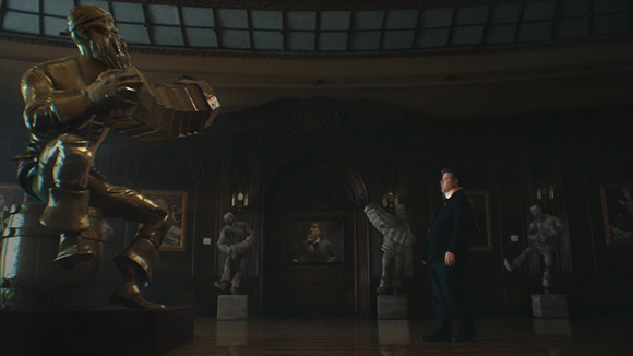 A man in a dark suit stands in a dim hall facing a large golden statue of a seated figure.