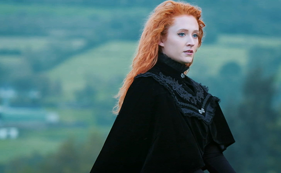 A woman with long wavy red hair and pale skin wearing a black velvet cloak against a blurred green landscape.