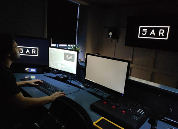 A person sits at a desk with three computer monitors and audio equipment in a dimly lit studio.