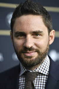 Close-up portrait of a man with dark hair and a beard wearing a checkered shirt and tie.