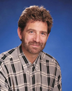 A man with wavy brown hair and a beard wearing a grey and black plaid button-down shirt against a blue background.