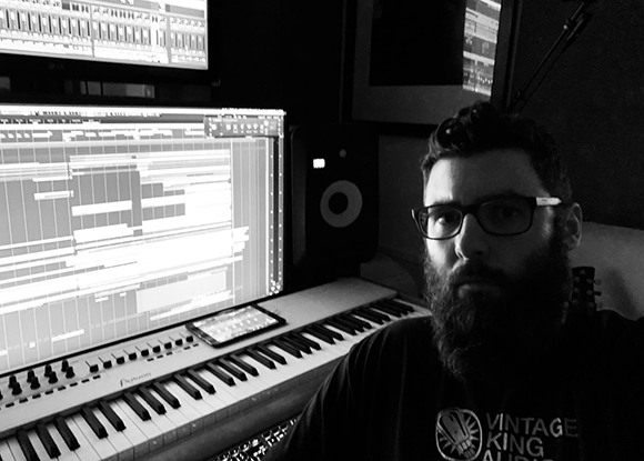 A bearded man wearing glasses sits in a music studio next to a keyboard and computer monitors.