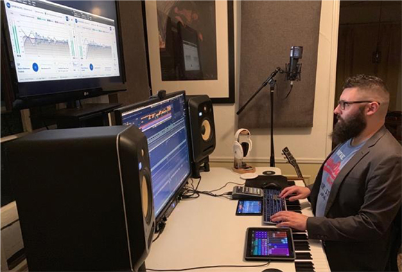 A bearded man in a suit uses a computer and keyboard in a recording studio with monitors and a microphone.