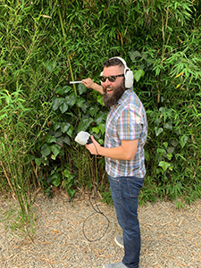 A bearded man wearing headphones and sunglasses holds a microphone and a directional recorder near bamboo.