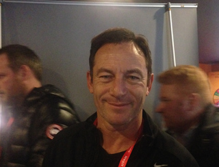 A man with short brown hair and a slight smile looks directly at the camera. He is wearing a black jacket.