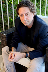 A man with wavy brown hair wearing a dark blazer and light trousers sits on a wicker chair.