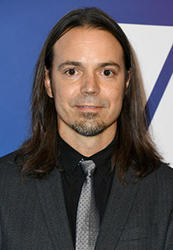 A man with long brown hair, a goatee, and a grey tie wearing a dark suit jacket.