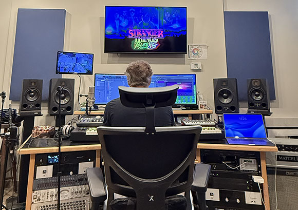 A person sits in a studio chair facing multiple computer monitors and studio speakers.