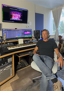 A man in a black t-shirt and jeans sits in a white chair in a music studio filled with recording equipment.