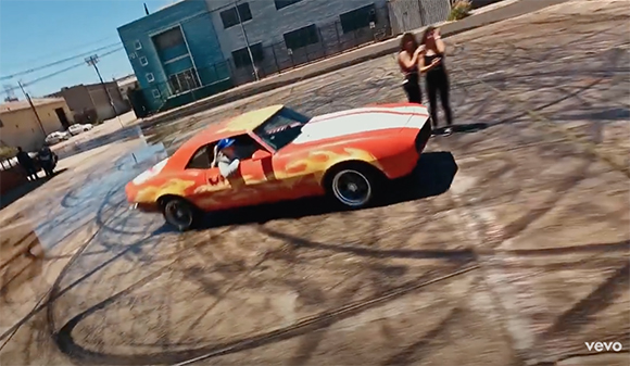 An orange muscle car with flame decals performs a drift in a paved lot with tire marks.