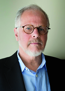 A man with gray hair, round glasses, and a white beard wearing a blue shirt and black blazer.