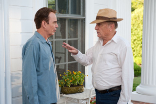 A man in a blue shirt faces an older man in a white shirt and tan hat on a porch.