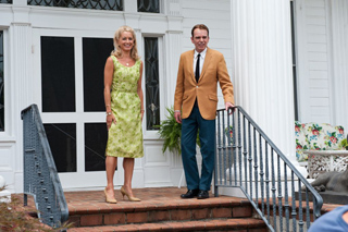 A woman in a lime green dress and a man in a tan blazer and blue trousers stand on white house steps.