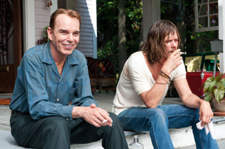 A man in a blue shirt smiles while sitting on steps next to a man in a white shirt smoking a cigarette.