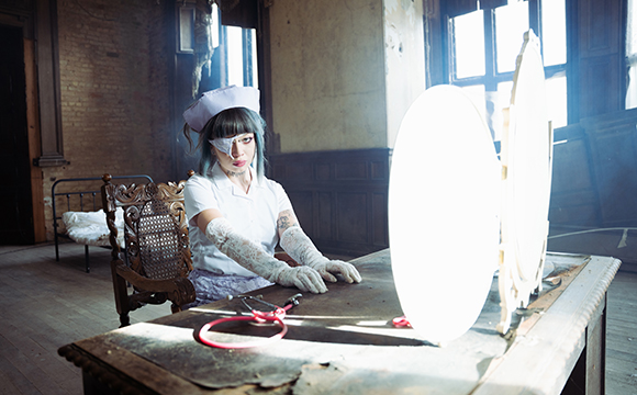 A person in a nurse outfit and eye patch sits at a wooden table in a dark, abandoned room.