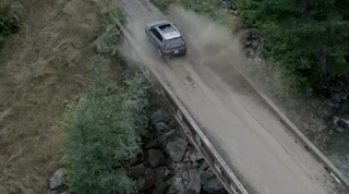 An aerial view of a silver SUV driving along a dirt road through a wooded area.