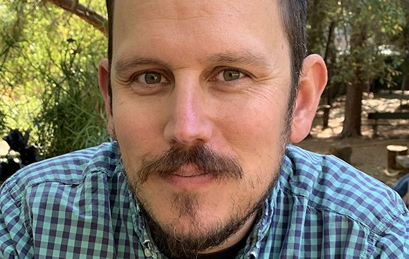 A man with short dark hair, a mustache, and a goatee wearing a blue and green checkered shirt.