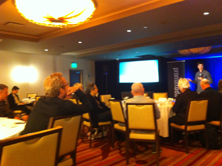 A speaker stands on a stage in front of a screen and banner, gesturing to an audience seated at round tables in a conference