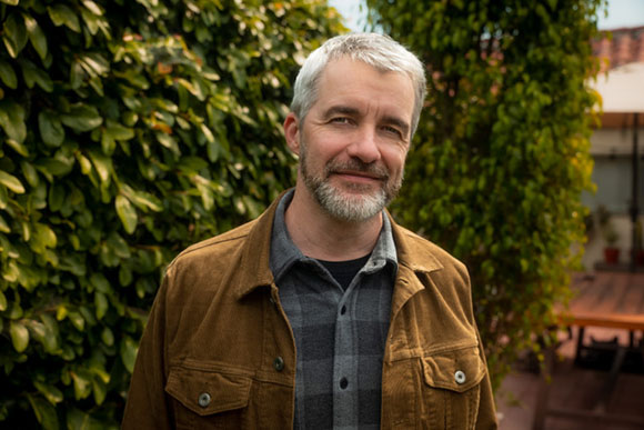 A middle-aged man with gray hair and a beard wearing a brown jacket and plaid shirt, smiling outdoors.