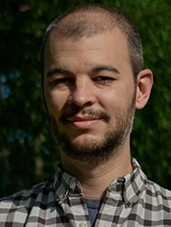 A man with short hair and a beard wearing a plaid shirt, smiling against a green foliage background.