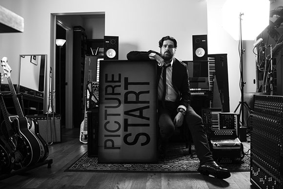 Black and white photo of a man sitting in a music studio next to a large sign that reads 'PICTURE START'.