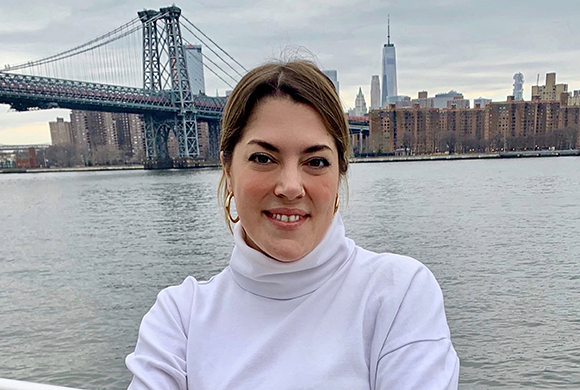 A woman in a white turtleneck smiling with the Manhattan Bridge and city skyline in the background.