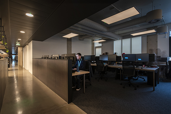 An open-plan office with desks, computer monitors, ergonomic chairs, and industrial ceiling pipes.