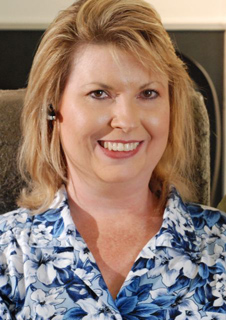 A smiling woman with blonde hair wearing a blue and white floral patterned shirt.