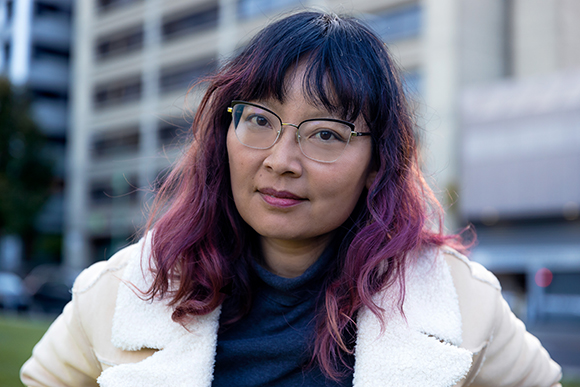 A woman with dark hair featuring purple tones and glasses looks forward against a blurred urban background.