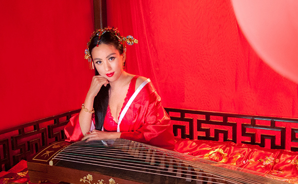 A woman in red traditional attire sits behind a guzheng against a red background.
