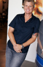 A smiling person wearing a dark blue short-sleeved shirt and grey jeans, leaning against a white wall.