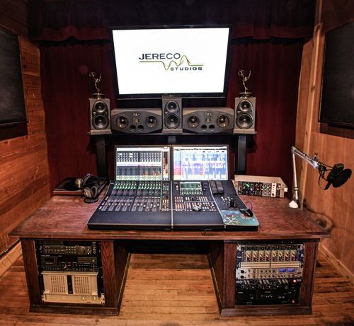 A large wooden desk holding a mixing console, computer monitors, studio monitors, and audio equipment.