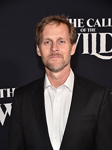 A man with light hair and a beard wearing a black suit and white button-down shirt.